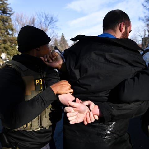 A person with their hands restrained behind their back, surrounded by people in vests labeled "police."