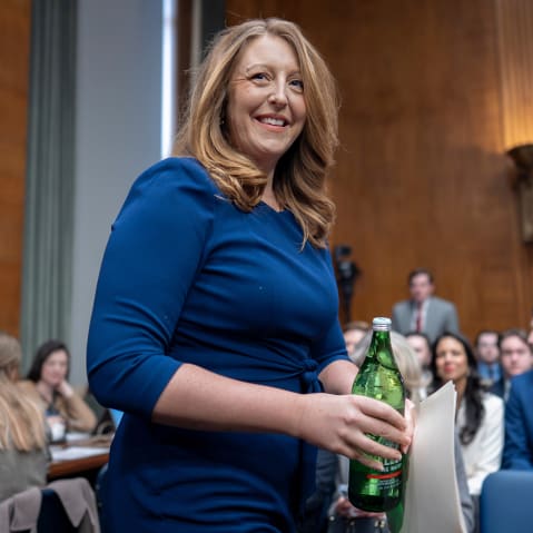 Casey Means is getting ready to sit down, as she is wearing a blue dress and holding a green bottle.