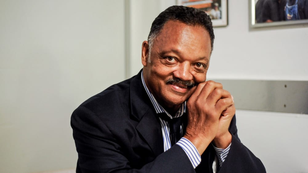 Rev. Jesse Jackson poses for a portrait, sitting down with his hands clasped up by his face.