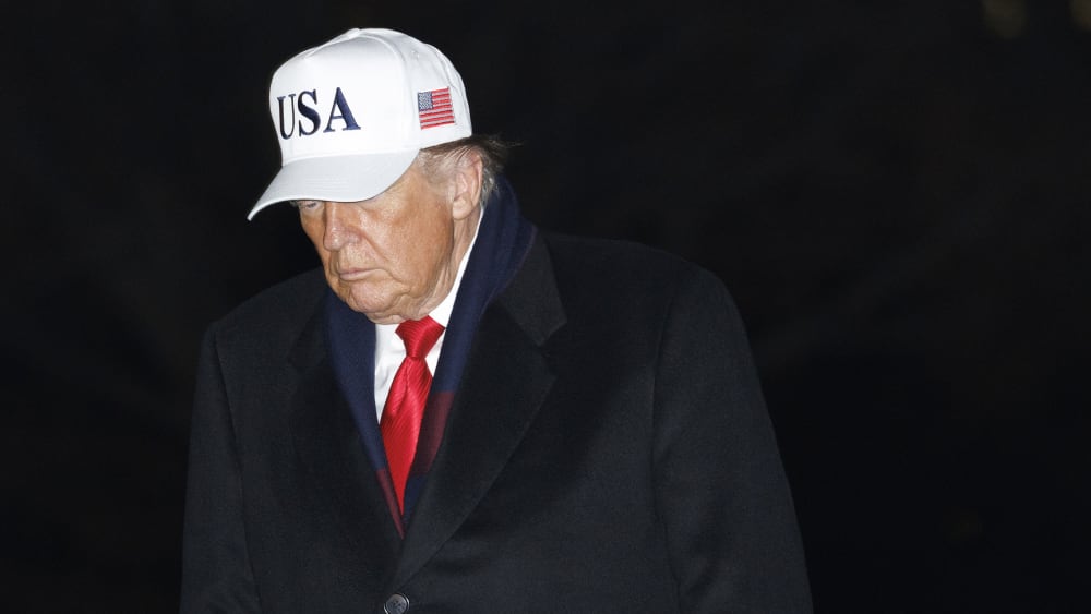 Donald Trump walks at the White House at night, wearing a white USA hat.