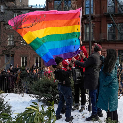 People raise a rainbow flag on a pole in a park. A crowd is gathered behind, watching and filming.