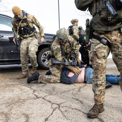 An ICE agent sits on top of a person, who is face down on the ground, and pins the person’s hands behind their back. Other agents stand nearby.