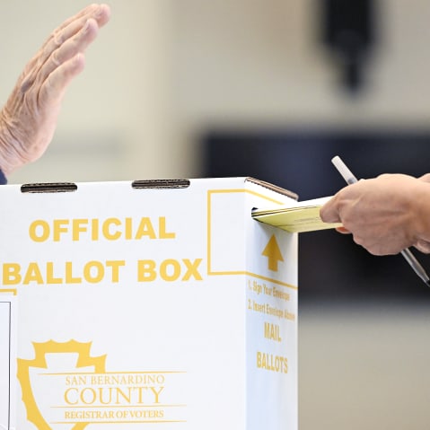 In a close-up shot, a person is putting their ballot in a box while an election official raises his hand.