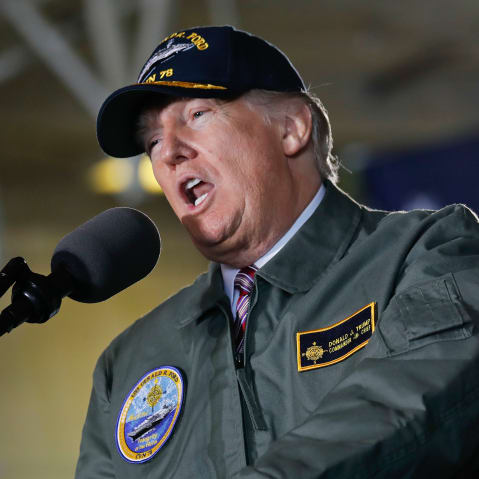 Donald Trump speaks into a microphone while wearing a USS Gerald R. Ford jacket and hat.