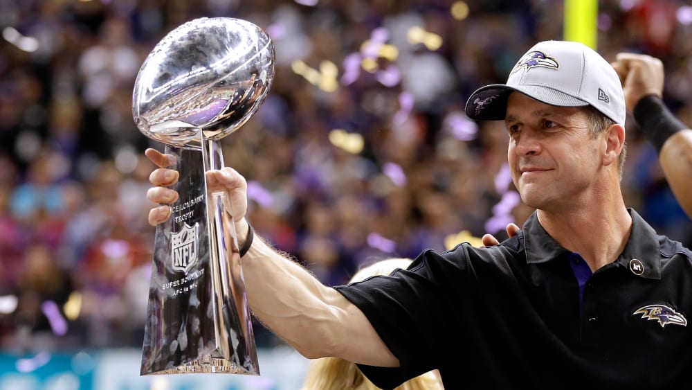 John Harbaugh holds the Vince Lombardi Championship trophy as purple and gold confetti falls in the background.