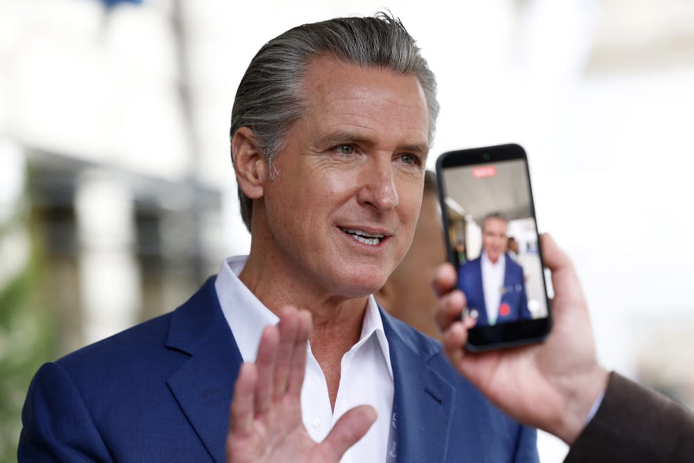 Gavin Newsom raises his right hadn as he speaks to journalists while one of them records a video of him.