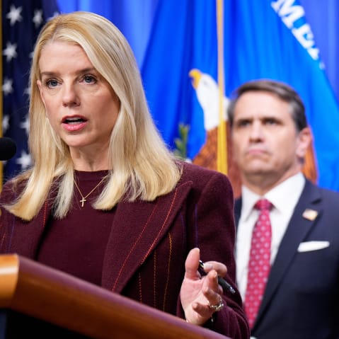 Pam Bondi stands behind a podium and microphone; Todd Blanche stands behind her, out of focus.