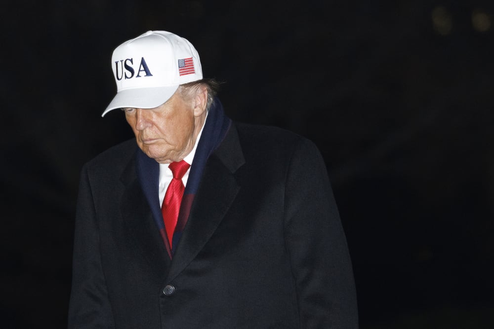 Donald Trump walks at the White House at night, wearing a white USA hat.