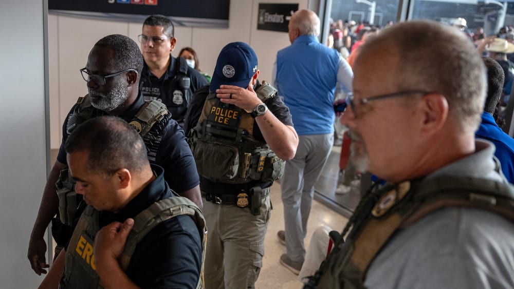 ICE agents stand in an airport. One hides their face by tipping their baseball cap down.