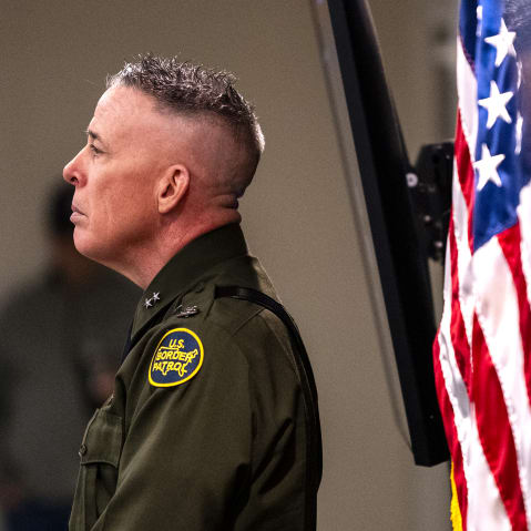 Greg Bovino is seen from the side while he looks on during a presser. His reflection can be seen behind him.