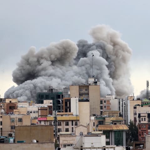 A great deal of smoke rises in the backdrop of a city skyline.