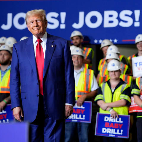 Donald Trump smirks in front of people in hard hats and yellow vests holding signs that say "DRILL BABY DRILL!" Behind them, a large poster that reads "JOBS! JOBS! JOBS!"