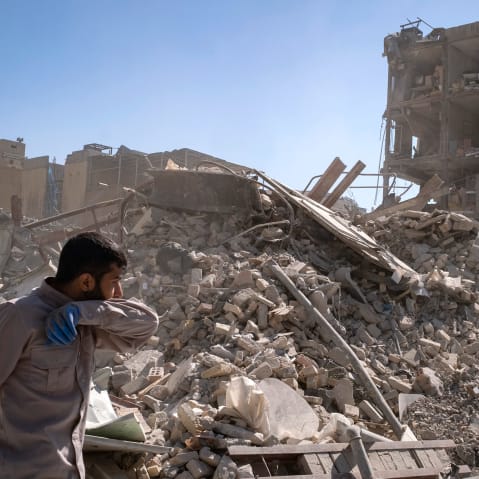 A person walks by a pile of rubble. Crumbling buildings are seen in the background.