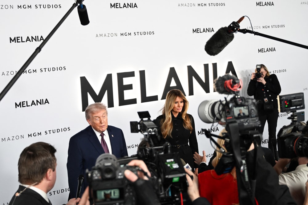 Donald and Melania Trump stand in front of a step-and-repeat banner advertising the movie "Melania." In front of them are journalists with cameras.
