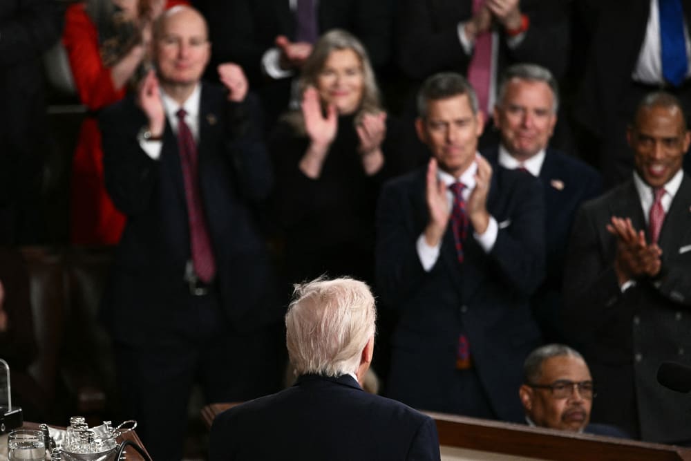 People clap, out of focus, as the back of Donald Trump's head is seen in focus in the foreground.
