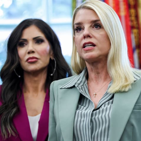 Kristi Noem is seen on the left side of the frame as she stands behind Pam Bondi. A small part of Donald Trump's face is visible on the bottom right.