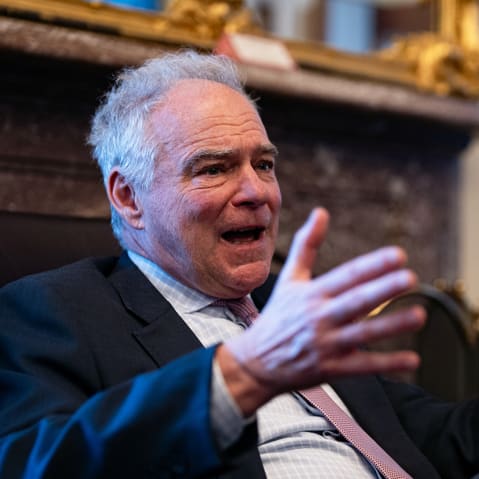 Tim Kaine, in focus on the left, gestures with his hands, while Rand Paul, out of focus on the right, looks at him.
