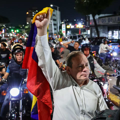 Juan Pablo Guanipa, center, stands in the street, holding a Venezuelan flag up with one hand. He is surrounded by people on motorcycles.
