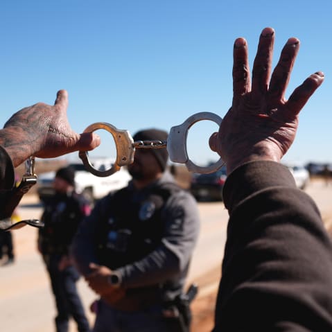 A close up photo shows a pair of hands holding handcuffs in the air. An officer can be seen in the background but out of focus.