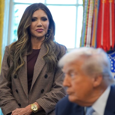 Kristi Noem, left, stands behind Donald Trump, out of focus in the foreground.