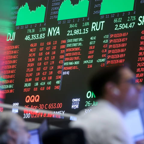 Stock exchange monitors can be seen in the background in focus as two men work in the foreground.