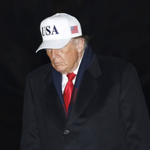 Donald Trump walks at the White House at night, wearing a white USA hat.