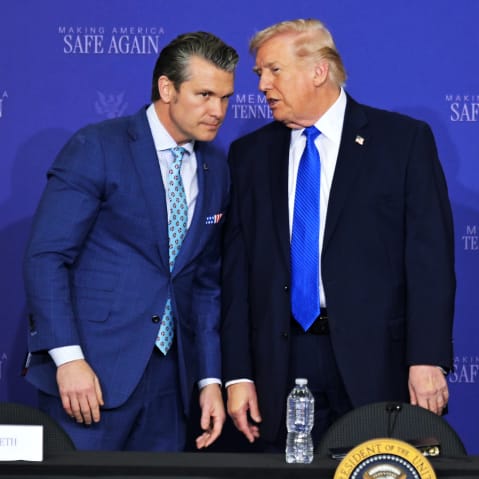 Pere Hegseth leans toward Donald Trump as Trump says something to his ear while they stand in front of a blue background.