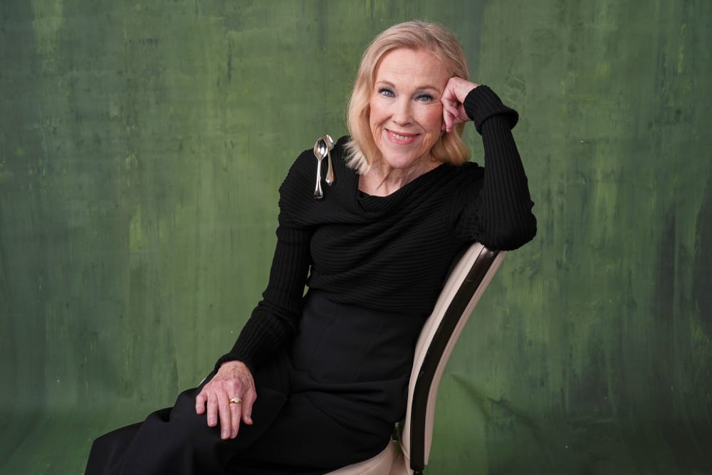 Catherine O'Hara in all black with two spoons on her shoulder. She sits in front of a painted green backdrop.