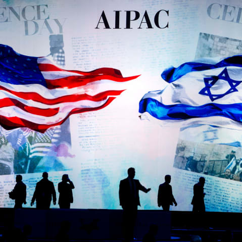 People are silhouetted before a screen that reads "AIPAC" and shows a visual of the American flag and the Israeli flag.
