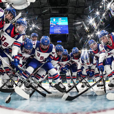 Hockey players in jerseys that read "USA" gather at the goal post. The photo is taken from a camera inside the goal.