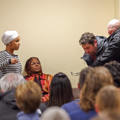 Ilhan Omar, left, raises her fist as if to throw a punch while security tackles a person from behind.