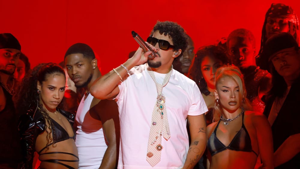 Bad Bunny, surrounded by performers, holds a microphone against a red background.