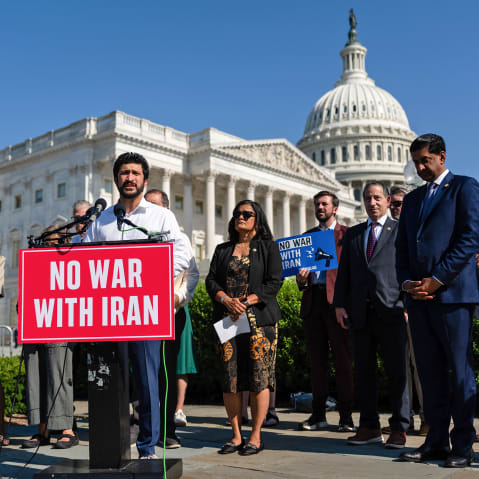Greg Casar stands behind a podium that has a red sign that says "NO WAR WITH IRAN." He is surrounded by other people, and in the background is the Capitol.