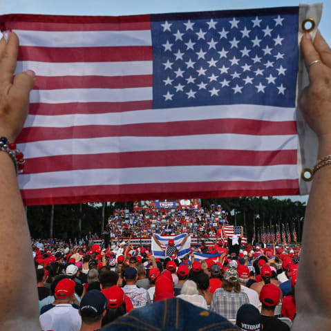 A person's arms are seen in the foreground holding an American flag while rally participants fill the rest of the frame in the background.