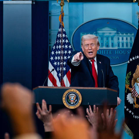 Donald Trump, in focus, stands a podium and points in front of him. Out of focus in the foreground, many people raise their hand.
