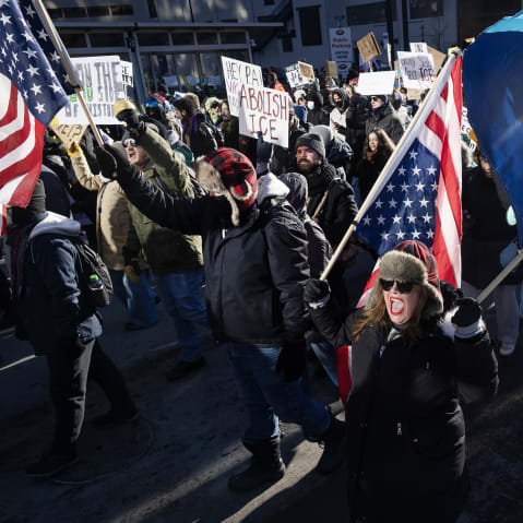 A female protestor shouts as she carries a flag in the foreground where others follow her during a daytime demonstration.