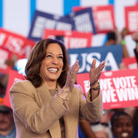 Kamala Harris claps; people behind her hold up signs in support.