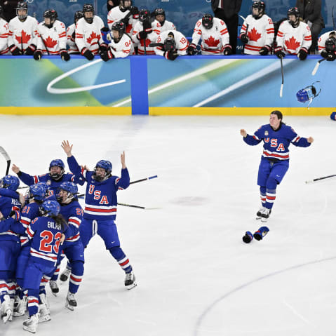 A group of US hockey players are huddled on the left side of the frame as one player runs toward them in celebration.