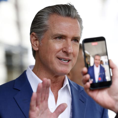 Gavin Newsom raises his right hadn as he speaks to journalists while one of them records a video of him.