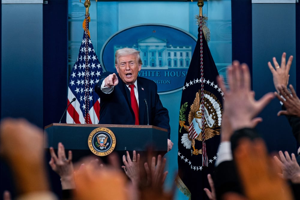Donald Trump, in focus, stands a podium and points in front of him. Out of focus in the foreground, many people raise their hand.