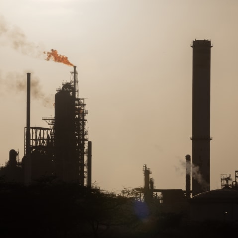 An oil refinery in Venezuela is seen in the distance while smoke and fire is coming out on the left side of the frame.