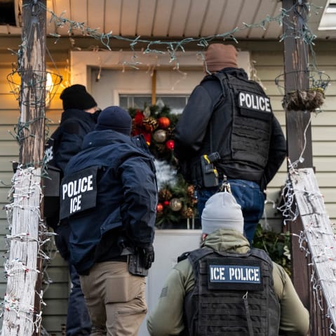 Four people with "police" and "ICE" on their uniforms knock on the door to a home decorated with a holiday wreath and string lights.
