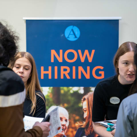 Two people talking with two job seekers in front of a “Now Hiring” sign.