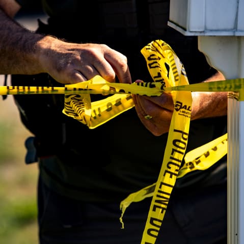 The hands of a police office tie yellow tape that says "Police Line Do Not Cross."