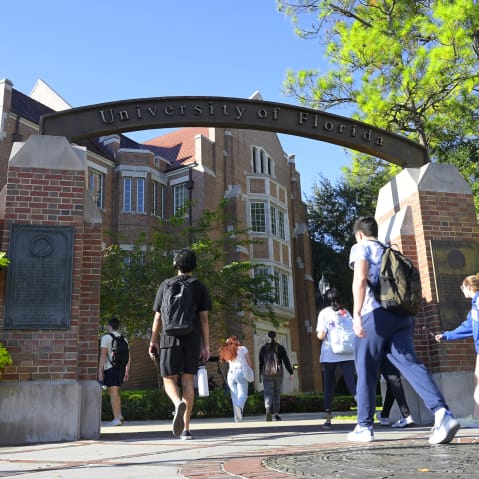 Students walk under the arch of University of Florida entrance on a sunny day.