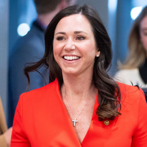 Katie Britt walks toward the camera with a smile on her face, wearing a red jacket.