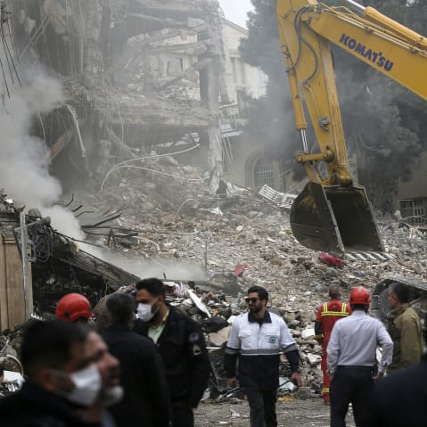 People are standing in front of a destroyed building as a work machine is seen in background.