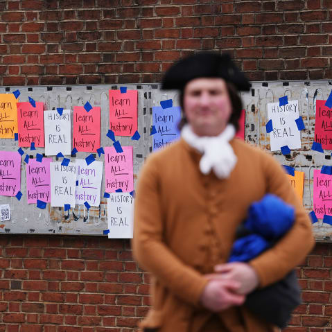 A person dressed in a late 18th- or early 19th- century costume stands blurred in the foreground. In focus in the background, multicolored paper signs that read "learn ALL history" and "HISTORY IS REAL" are taped to an empty panel on a brick wall.