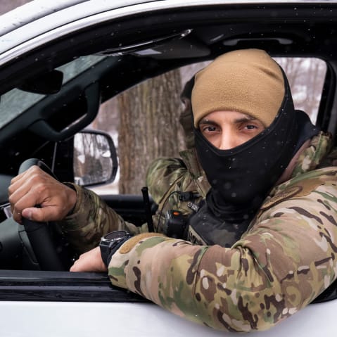 A masked agent driving a white car looks out the window of the car.