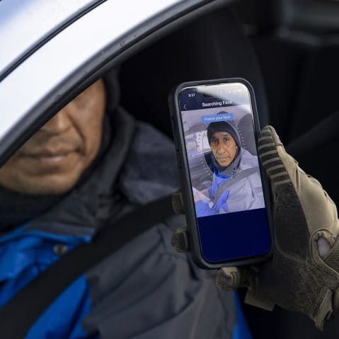 A federal agent holds a phone up to a person in the driver's seat of a car. The phone screen shows a program that is scanning the driver's face.